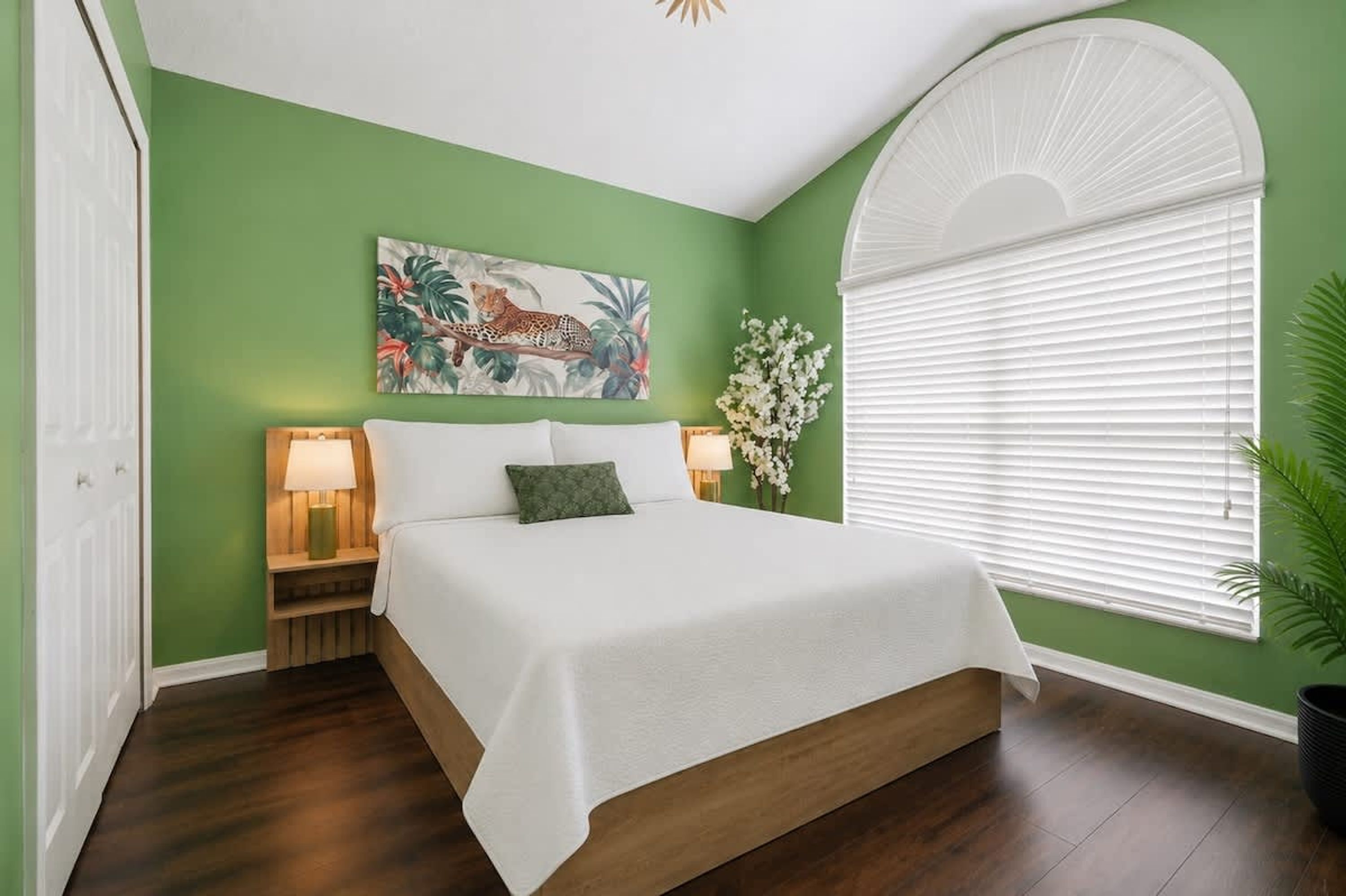 A bedroom with a bed, green walls, a window with blinds, a nightstand with a lamp, and a painting on the wall.