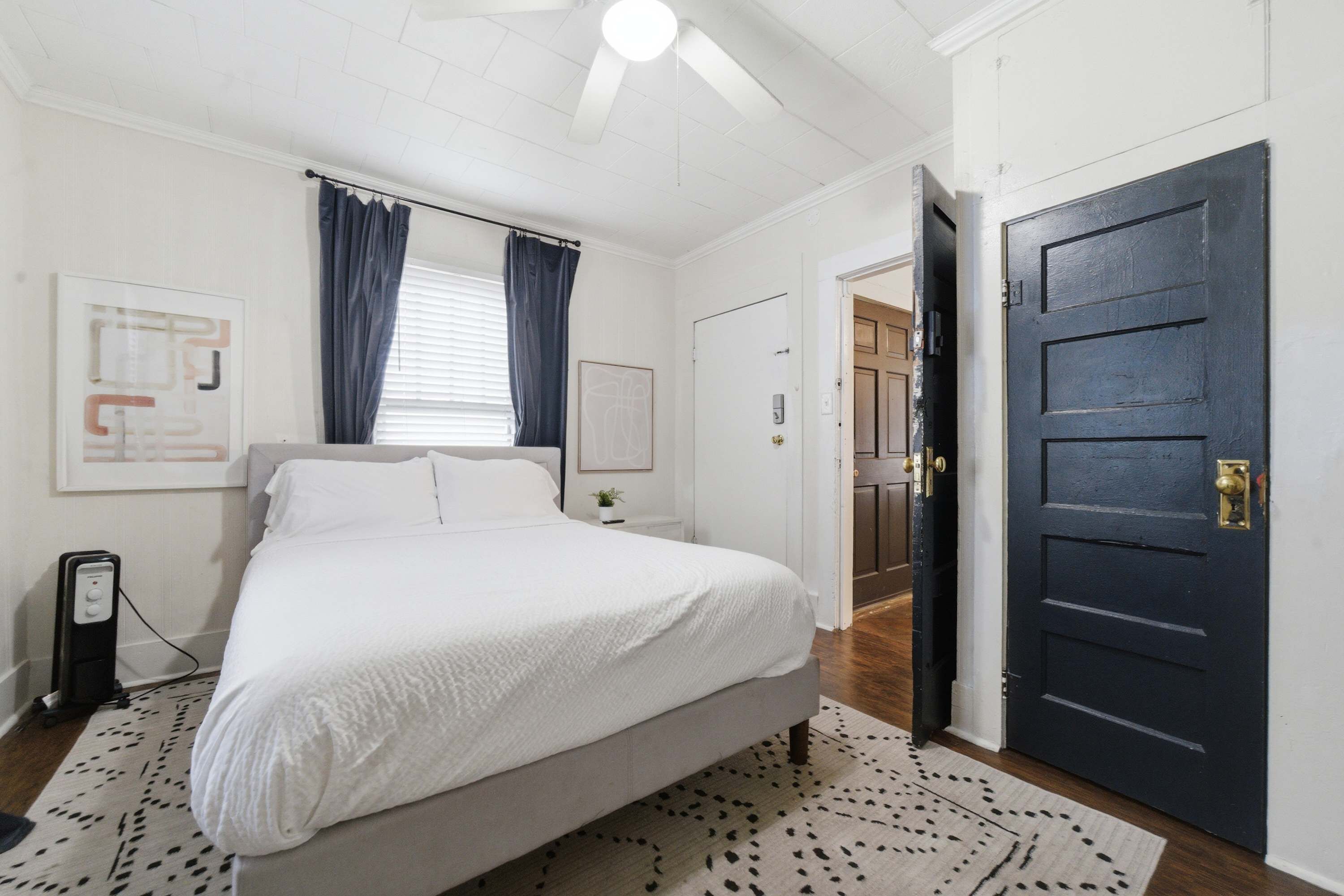 A bedroom with a bed, a nightstand, a wall-mounted heater, a door, and a window with curtains.