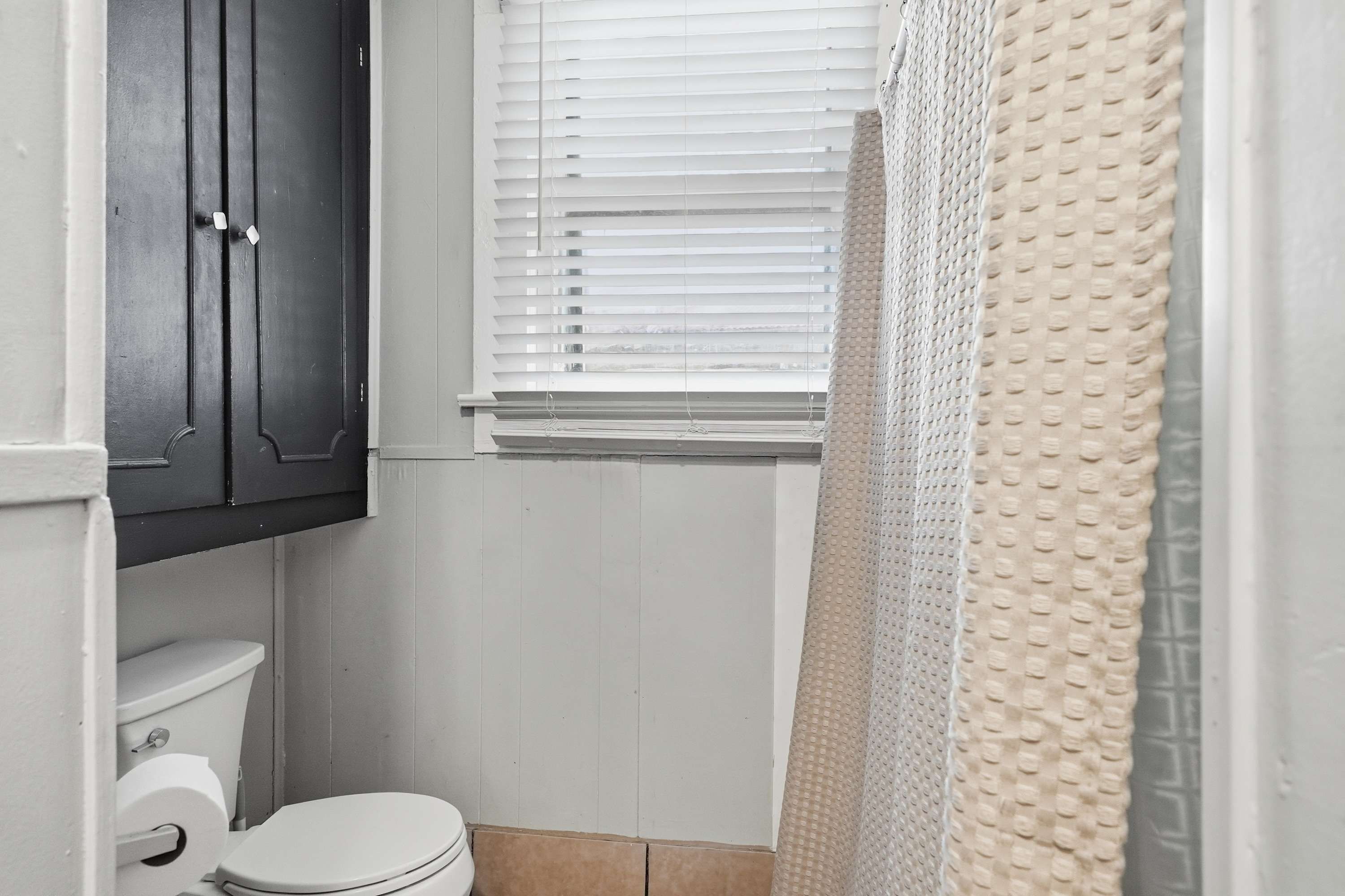 Une salle de bain avec des toilettes, un meuble en bois foncé, des murs blancs et une fenêtre avec des stores.