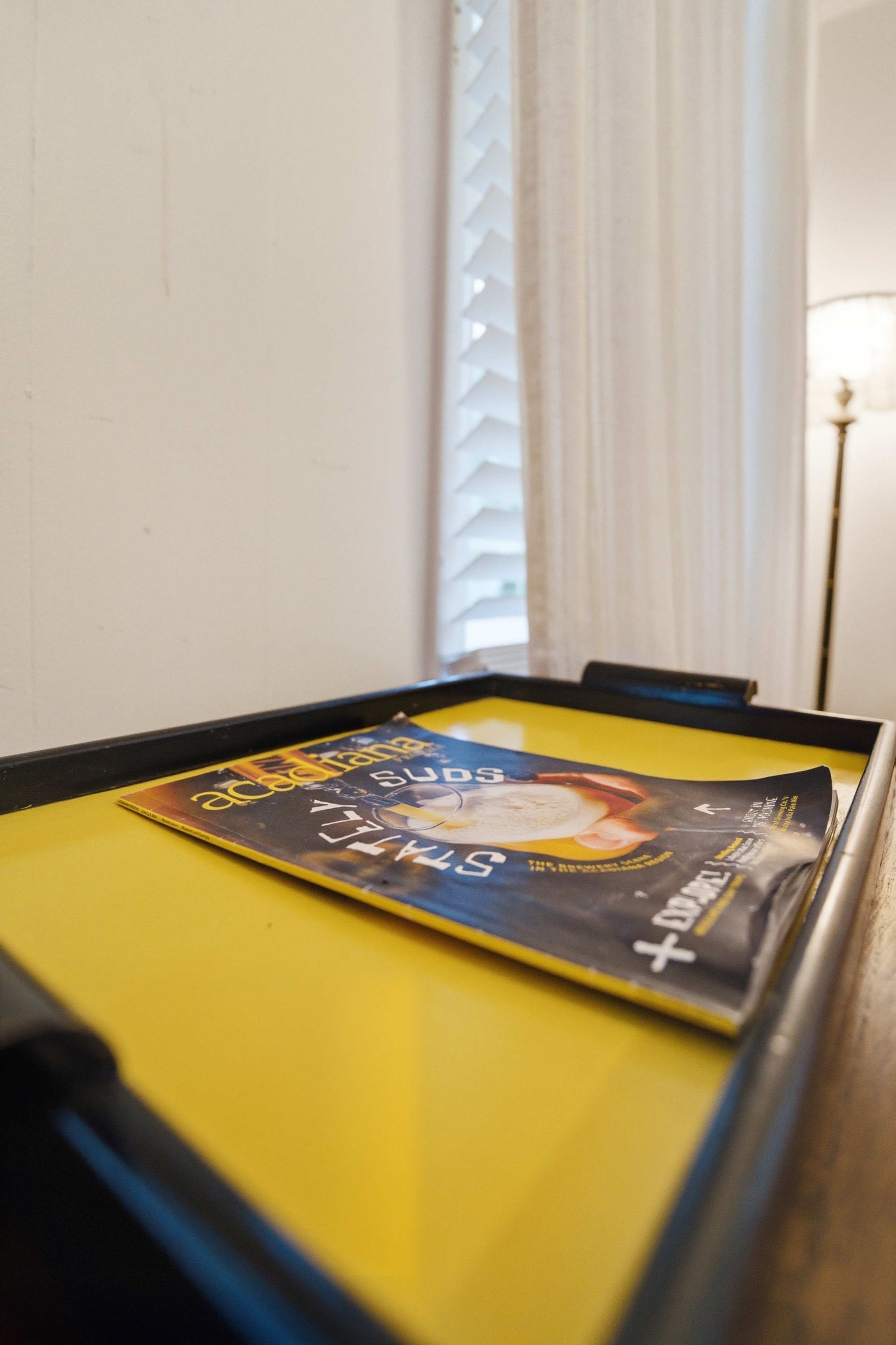 A table with a magazine on it, a window with blinds, and a lamp in the background.