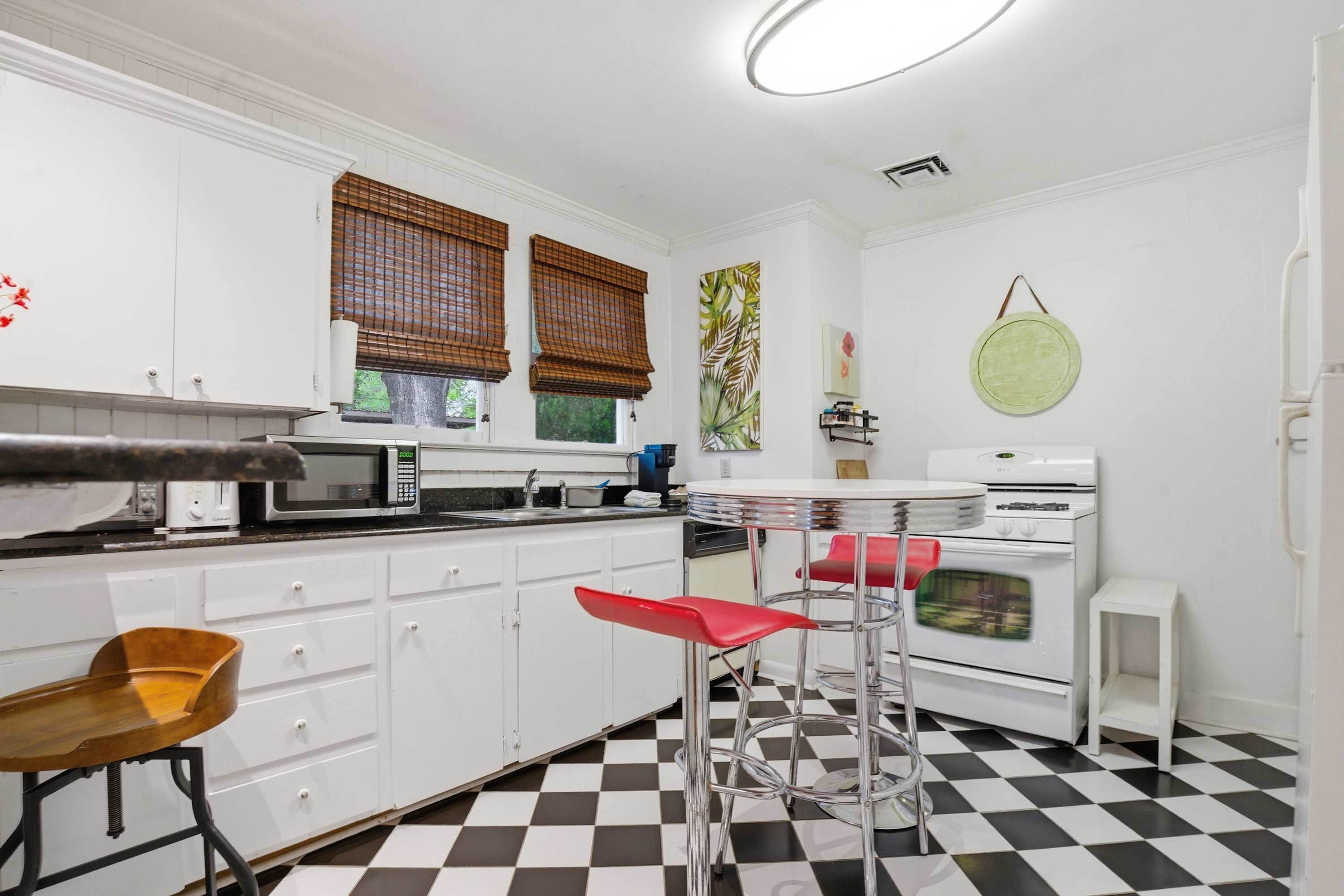 A compact kitchen with white cabinets, a microwave, a stove, and a sink. There are two stools and a small table with a round top.