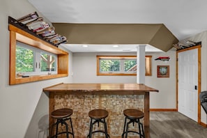 Zona de bar con mostrador de madera y taburetes, dos ventanas con vistas a follaje y una puerta con una placa.