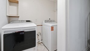 A laundry room with a front-loading washer and dryer, a wooden shelf, and a white wall.