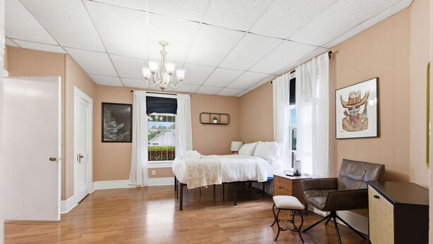 A hotel room with a wooden floor, a bed with white linens, a desk with a chair, a chandelier, and a window with curtains.