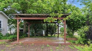 A covered patio area with a wooden pergola, brick flooring, and surrounding greenery.
