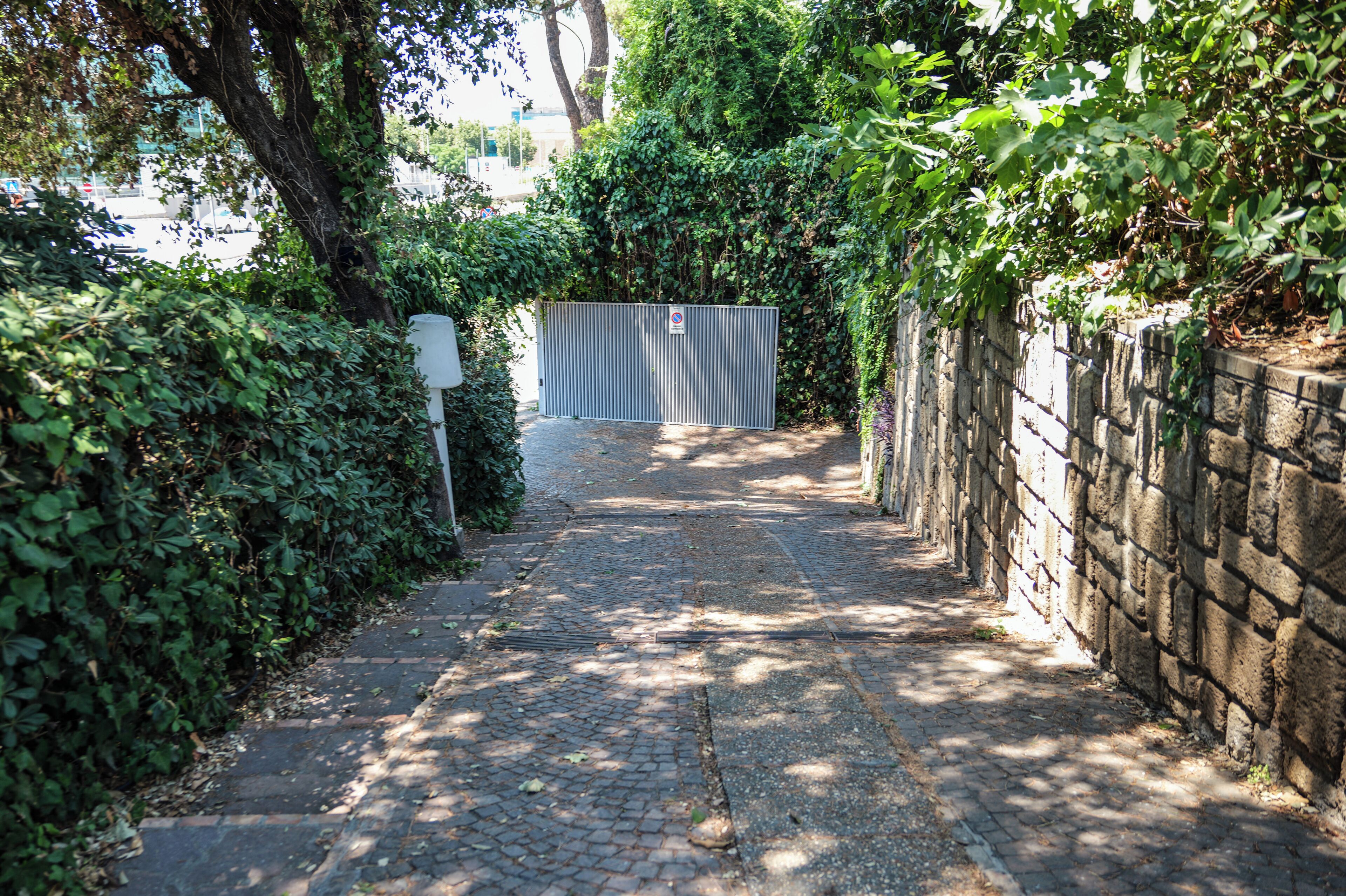 A paved pathway leading to a gated entrance with a metal fence and a sign.