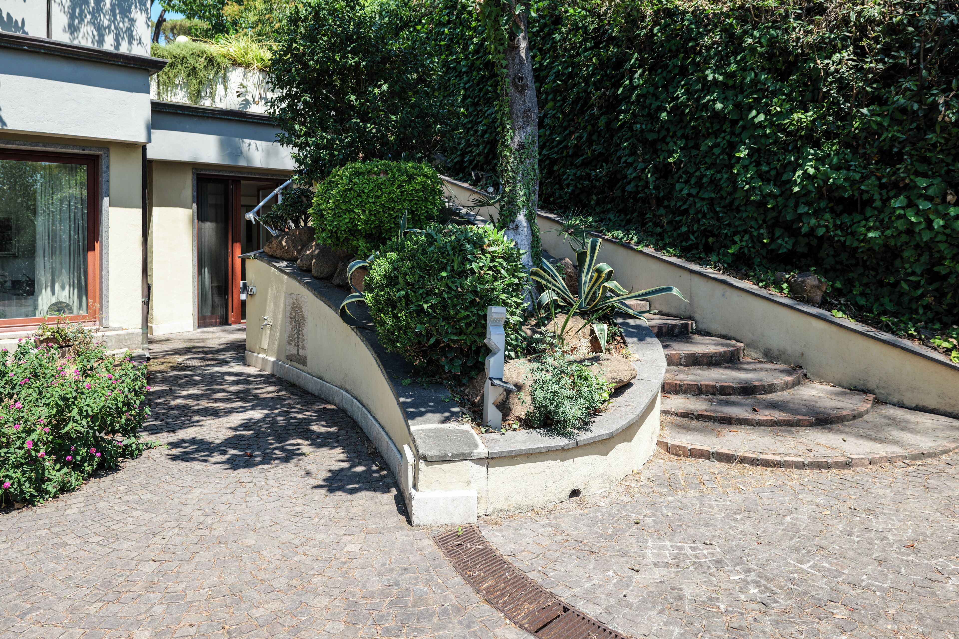 Un chemin pavé mène à un bâtiment doté d’une entrée incurvée, entouré de verdure et d’un petit espace aménagé en jardin.