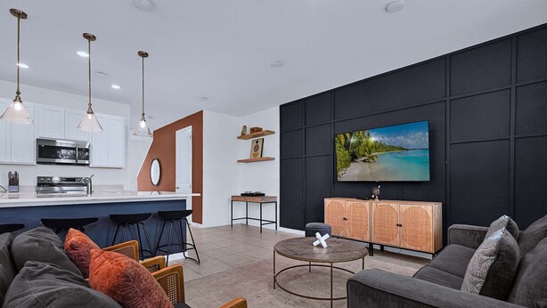 Uma sala de estar moderna com uma televisão de tela plana grande, uma parede de destaque escura e uma área de cozinha com armários brancos e um balcão.