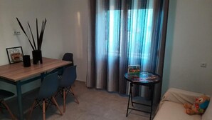 Un área de comedor con una mesa de madera y sillas, una mesita redonda con un juego de mesa y un peluche en un sofá.