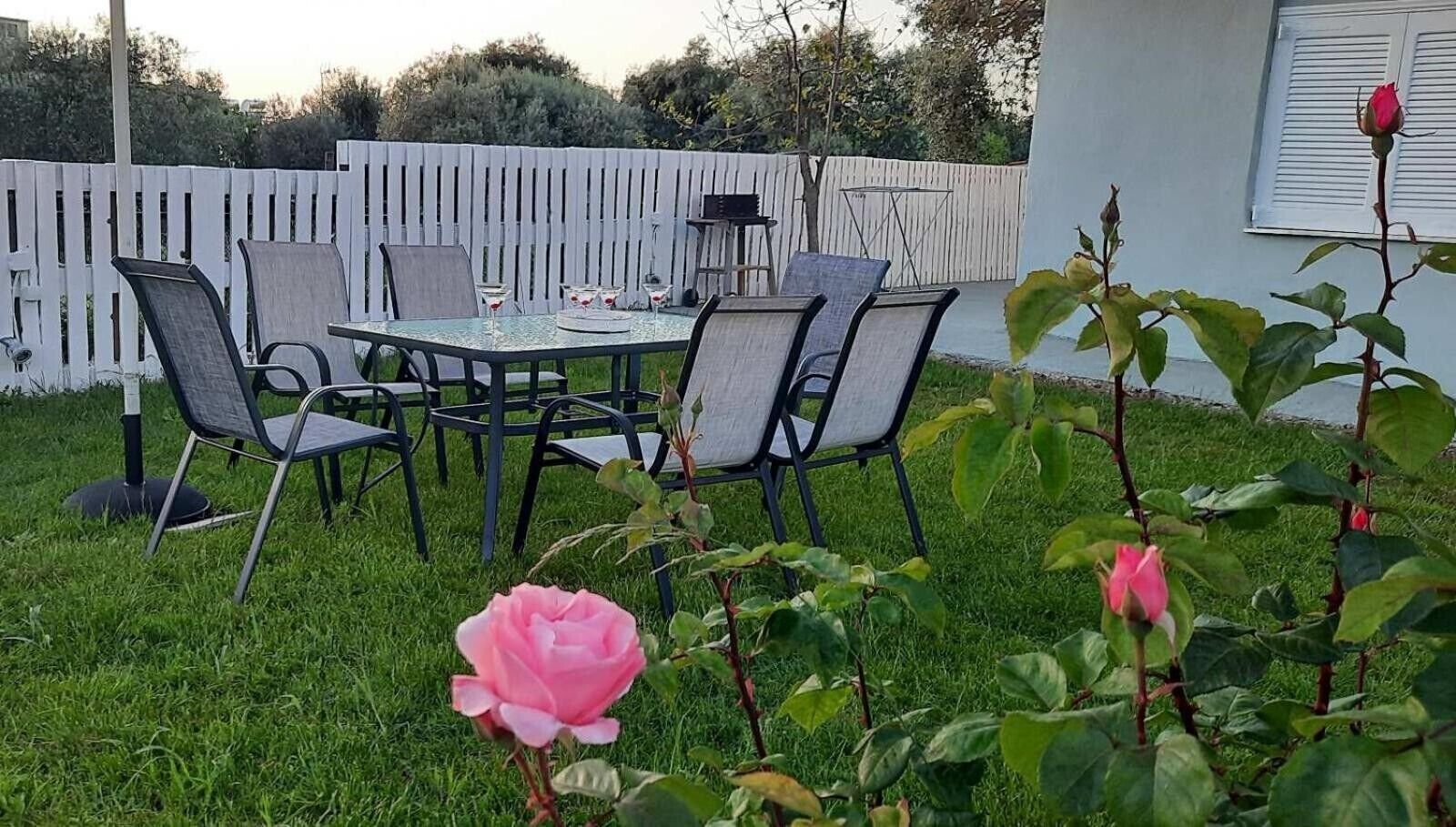 A patio with a table and chairs, surrounded by a white fence and greenery.
