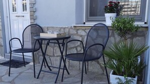 A small outdoor seating area with a table and two chairs, a potted plant, and a window with a view. - Apartment 'Chrissi Princess 1' with Private Garden, Wi-Fi and Air Conditioning (Skala Sotiros)