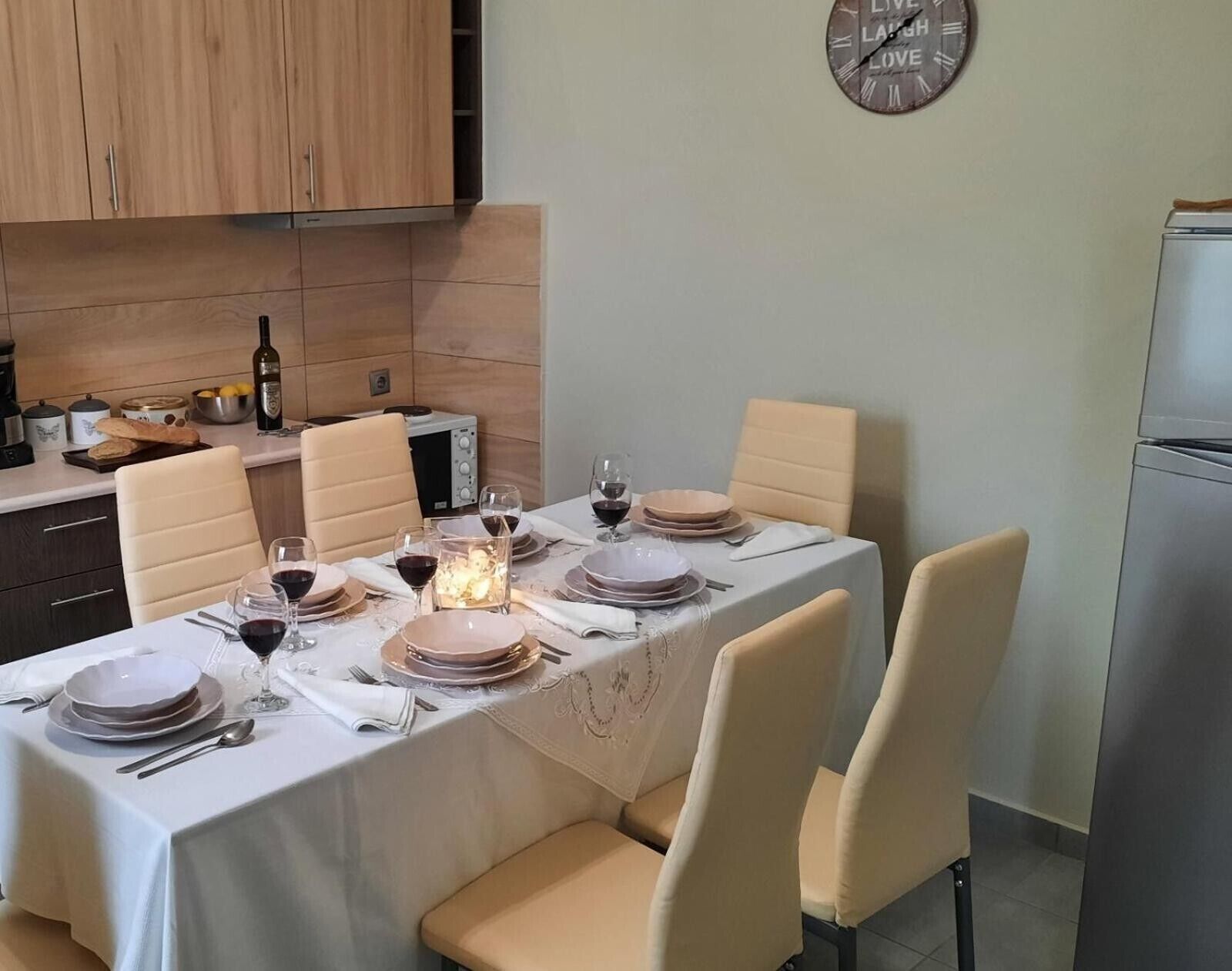 Une salle à manger avec une table dressée pour six personnes, une horloge au mur et une cuisine en arrière-plan.