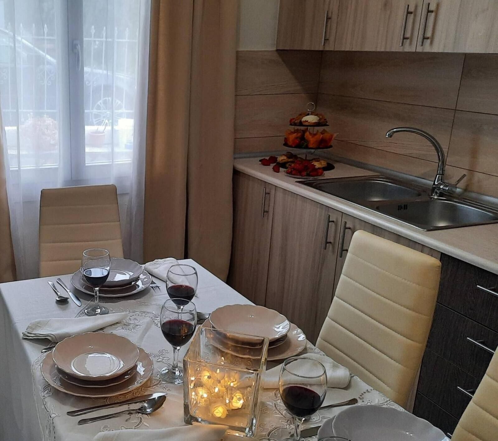 Un comedor con una mesa preparada para dos, una cocina con un fregadero y un ventanal con cortinas.