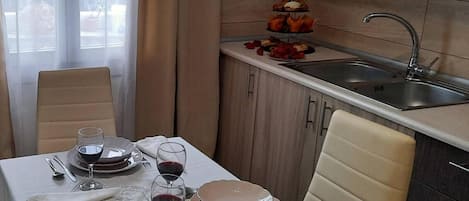 Un coin repas avec une table dressée pour deux personnes, une cuisine avec un évier et une fenêtre avec des rideaux.