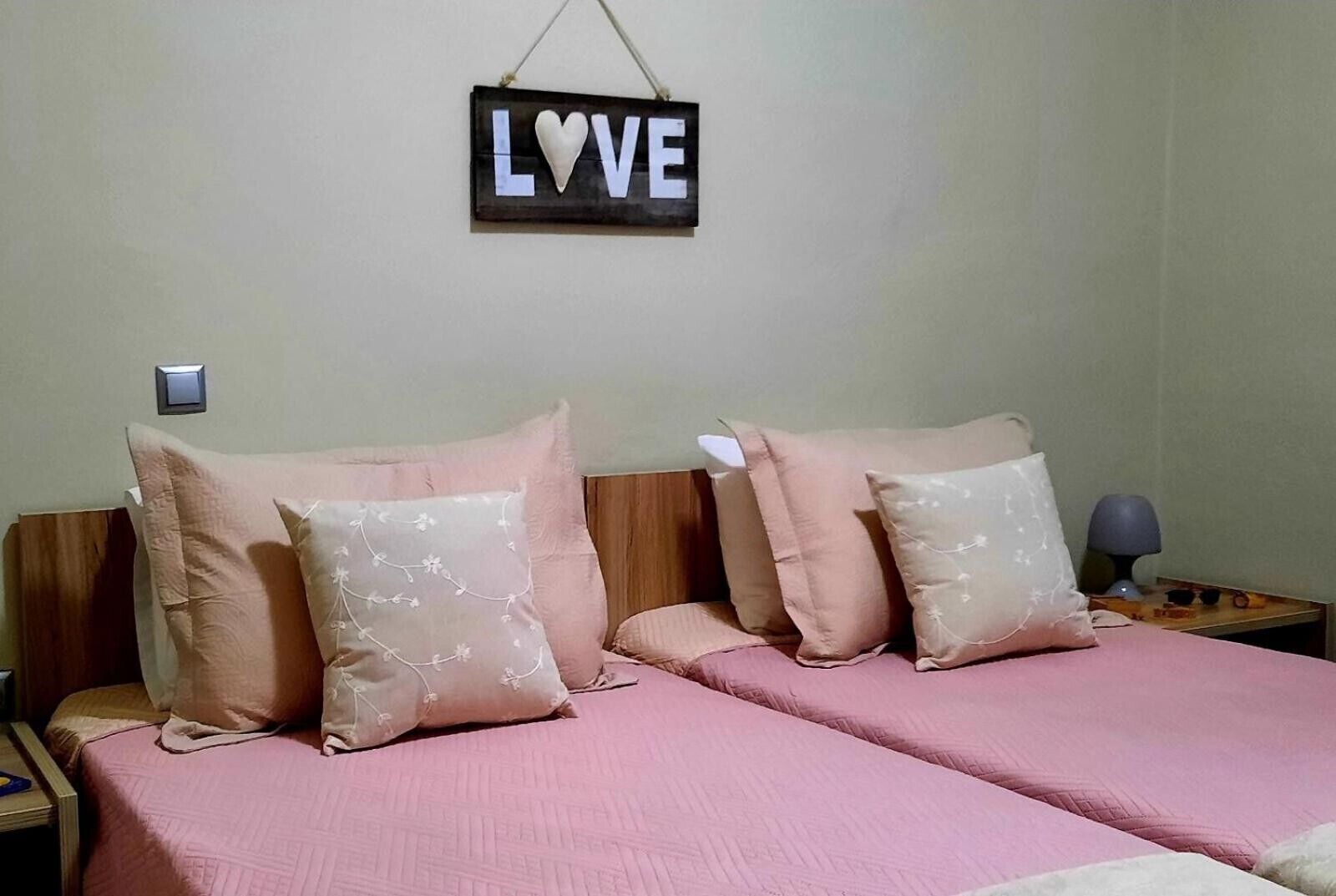 Une chambre avec deux lits, chacun agrémenté de draps roses et de coussins décoratifs. Un panneau portant le mot « AMOUR » est accroché au-dessus des lits.