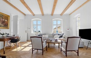 Amplia sala de estar con un sofá grande, dos sillones, una mesa de centro y un televisor. Se observan dos ventanas con arcos, vigas de madera en el techo y un cuadro en la pared.