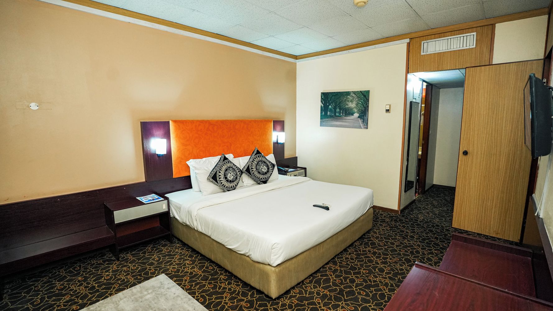 A hotel room with a bed, nightstands, a wooden wardrobe, a painting on the wall, and a patterned carpet.