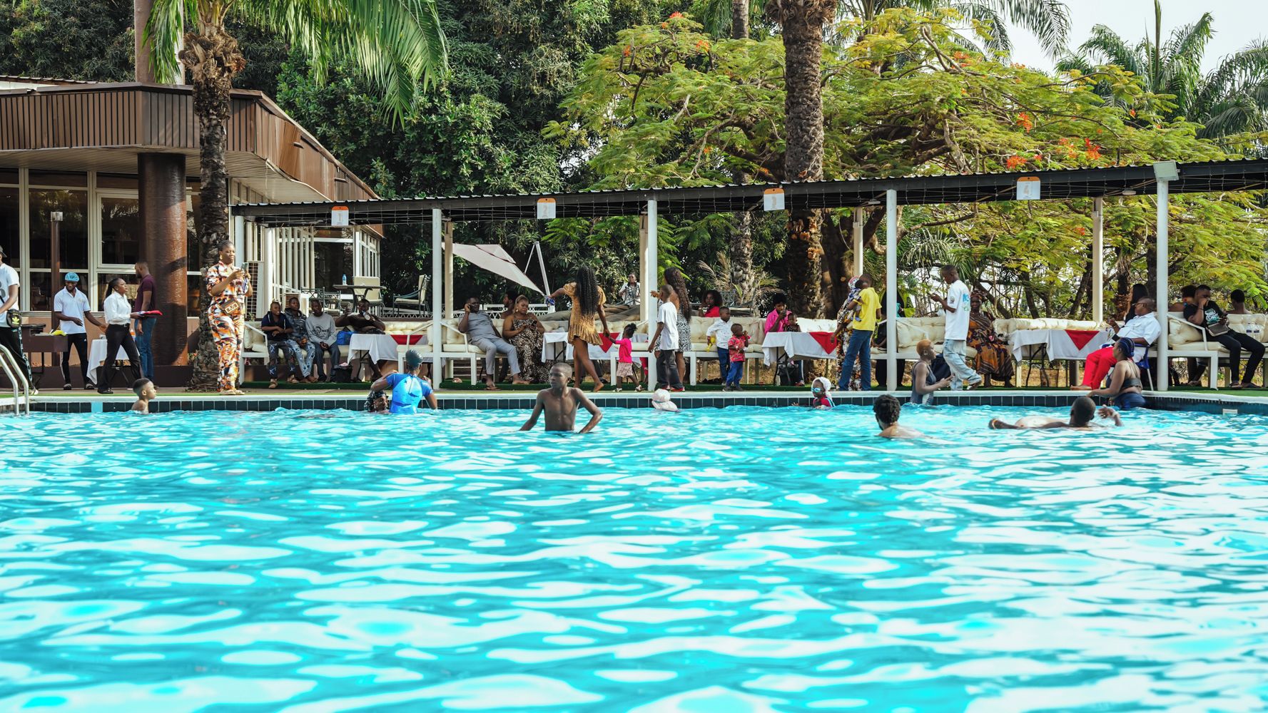 Piscina com pessoas ao redor, área coberta com mesas e cadeiras, e um edifício ao fundo.