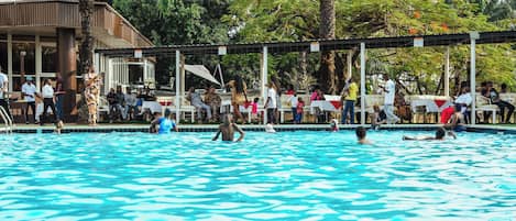 A swimming pool with people around it, a covered area with tables and chairs, and a building in the background.