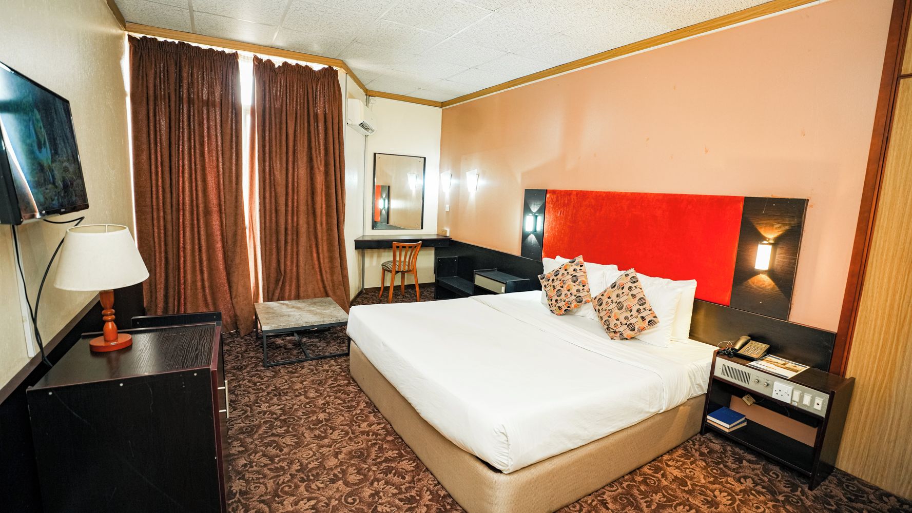 A hotel room with a bed, a desk, a chair, a TV, and a nightstand with a lamp and phone.