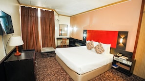 A hotel room with a bed, a desk, a chair, a TV, and a nightstand with a lamp and phone. - Landmark Nike Lake Resort (Enugu)