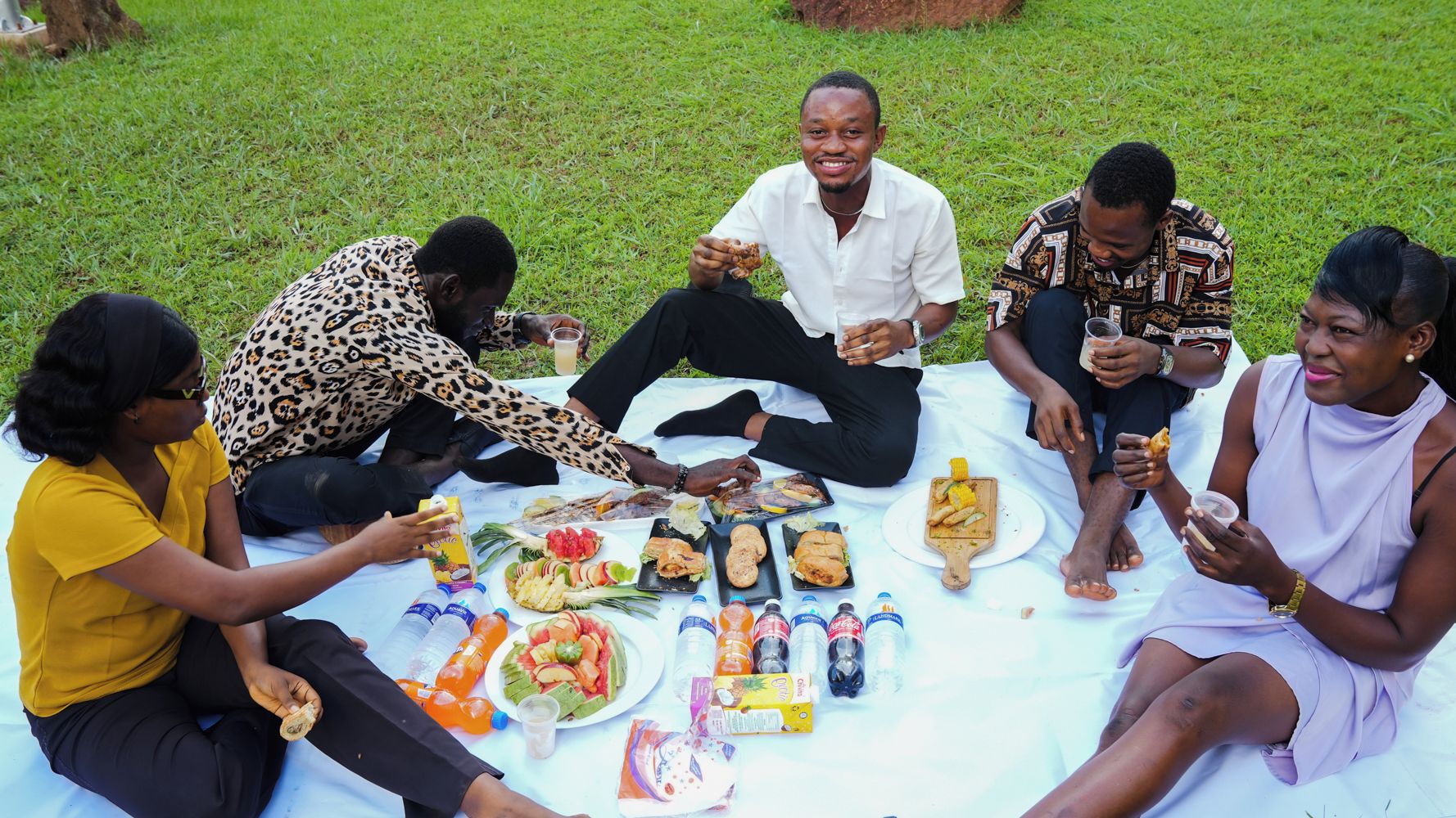 A group of people sitting on a blanket outdoors, enjoying a meal with various food items and drinks.