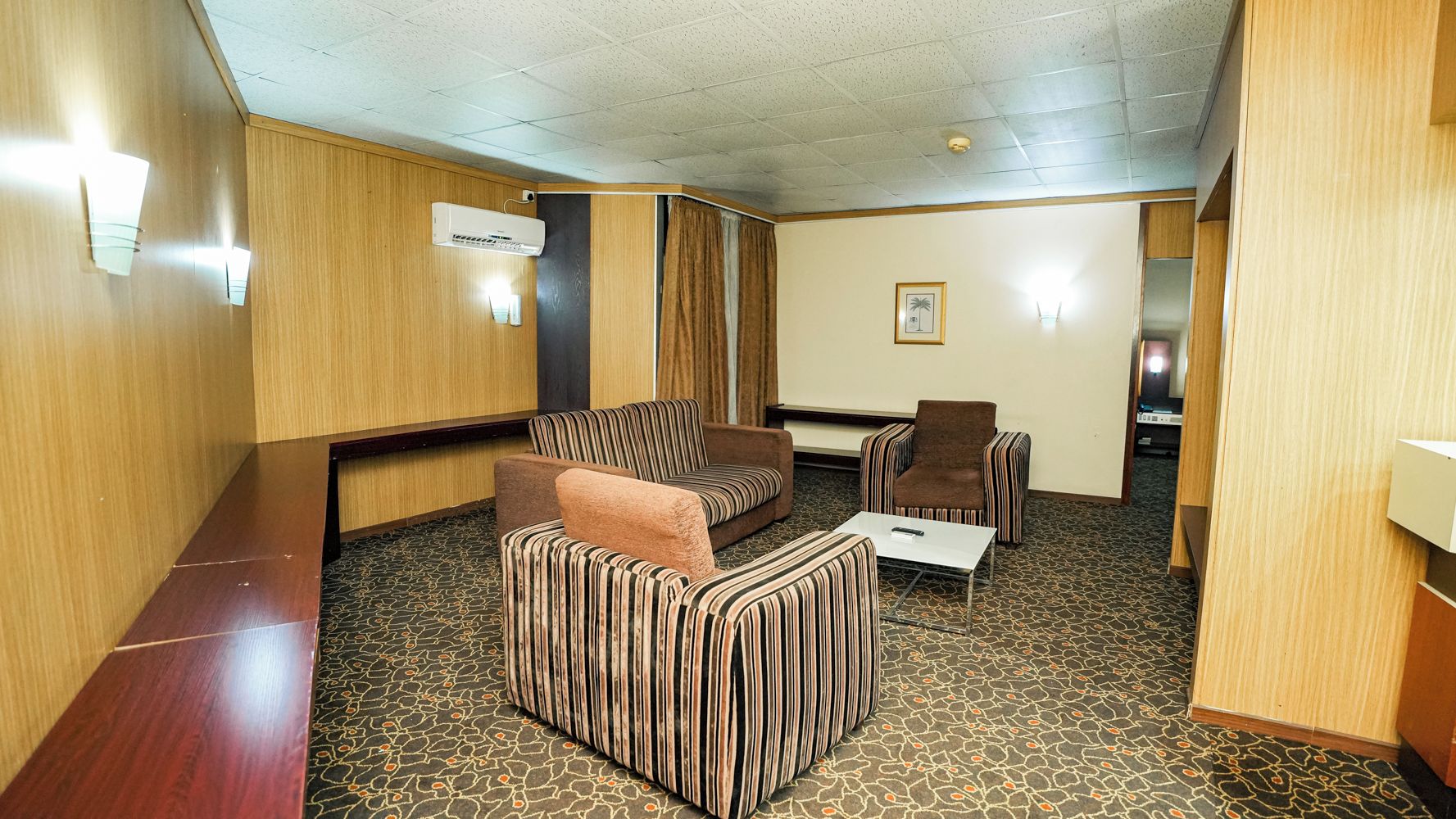 A hotel room with a wooden panel wall, a patterned carpet, a sofa, a chair, and a small table.