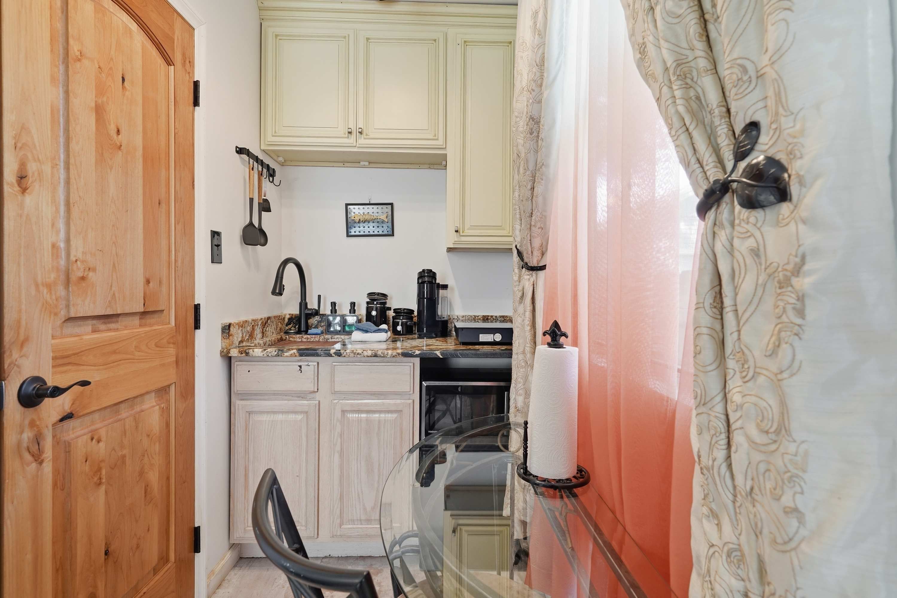 A compact kitchen with wooden cabinets, a countertop with various appliances, and a glass door leading to a bathroom.