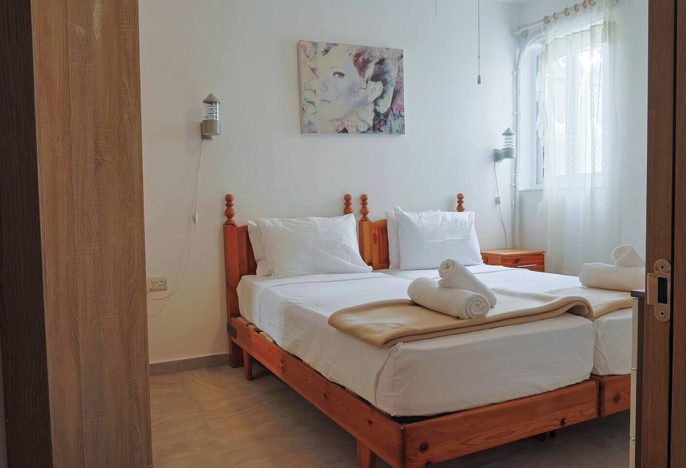 Uma cama de madeira com roupa de cama branca, uma mesinha de cabeceira, uma luminária na parede e um quadro acima da cama.