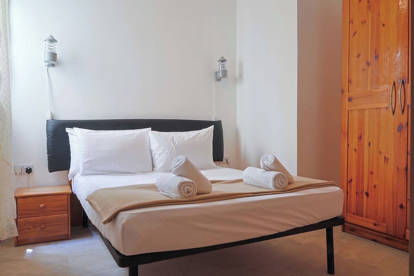 A neatly made bed with white linens and rolled towels, a wooden nightstand with a drawer, and a wooden wardrobe.