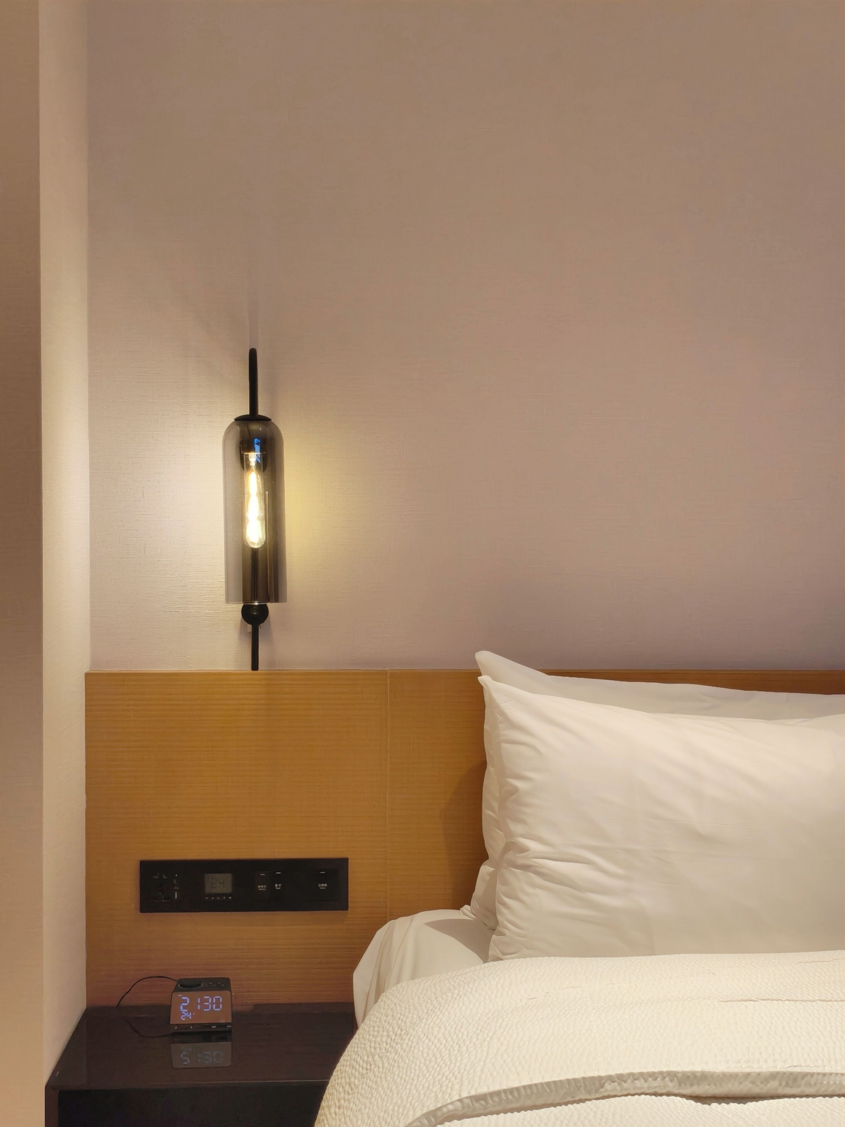 A hotel room with a bed, a nightstand with a digital clock, and a wall-mounted light fixture.