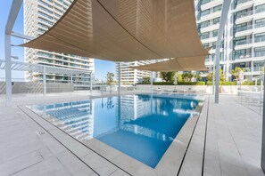 Ein Schwimmbecken mit glasklarer, blauer Wasseroberfläche, umgeben von einer weißen Fliesenveranda und beschattet durch große, beige farbene Sonnensegel.