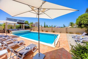 Ein Pool mit Liegestühlen und Sonnenschirmen, umgeben von einer gefliesten Terrasse und viel Grün.