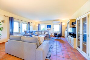 A living room with a sofa, a coffee table, and a TV. There are windows with curtains, a lamp, and a cabinet with glass doors. - Casa Serrao | Carvoeiro | Private Pool & Sea Glimpses | Walk to Dining & Beaches (Carvoeiro)