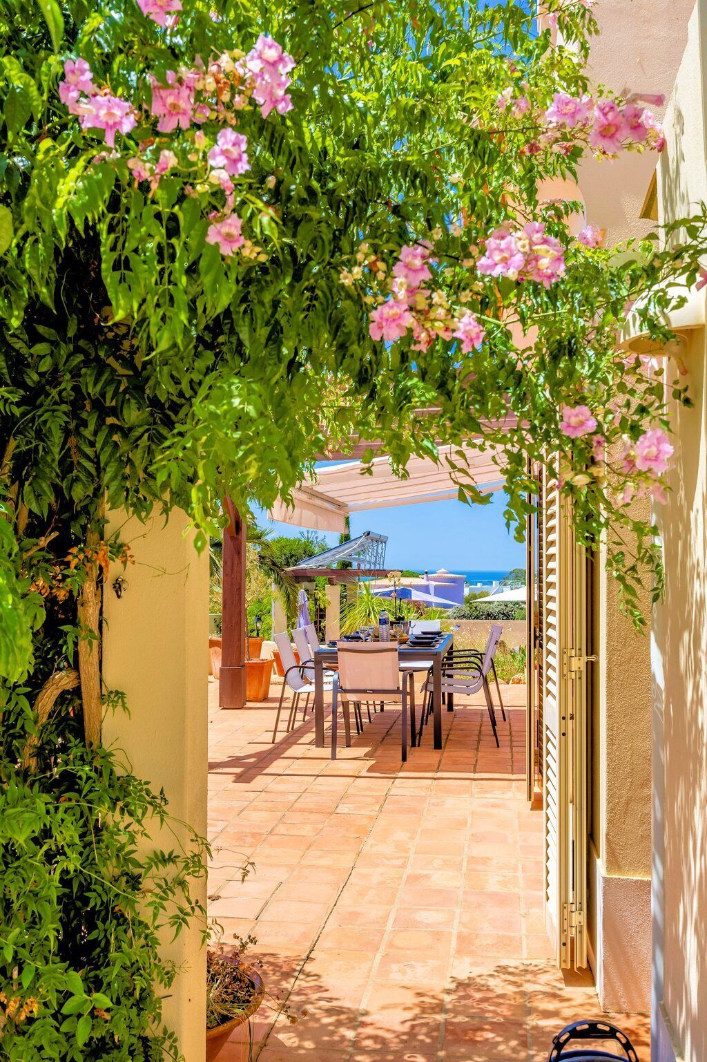 An outdoor patio with a table and chairs, surrounded by greenery and pink flowers.