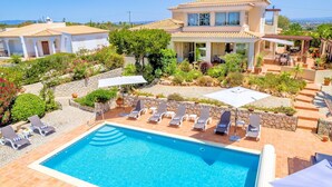Uma piscina com espreguiçadeiras e guarda-sóis, cercada por um jardim bem cuidado e uma casa de dois andares.