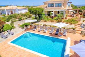 A swimming pool with lounge chairs and umbrellas, surrounded by a landscaped garden and a two-story house. - Casa Serrao | Carvoeiro | Private Pool & Sea Glimpses | Walk to Dining & Beaches (Carvoeiro)