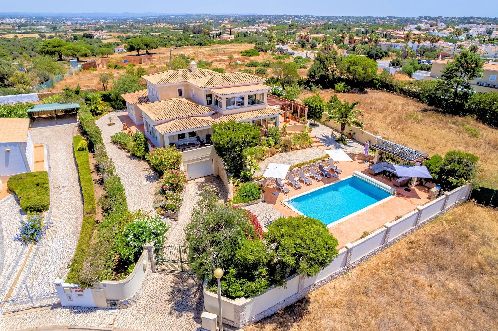 A two-story house with a swimming pool, surrounded by a garden and a view of a residential area.