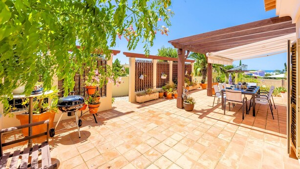 Um pátio externo com churrasqueira, plantas em vasos, pérgola e mesa com cadeiras.