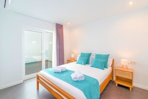 Une chambre d’hôtel moderne avec un grand lit, deux tables de chevet, une porte-fenêtre coulissante donnant sur un balcon et un design simple et épuré.