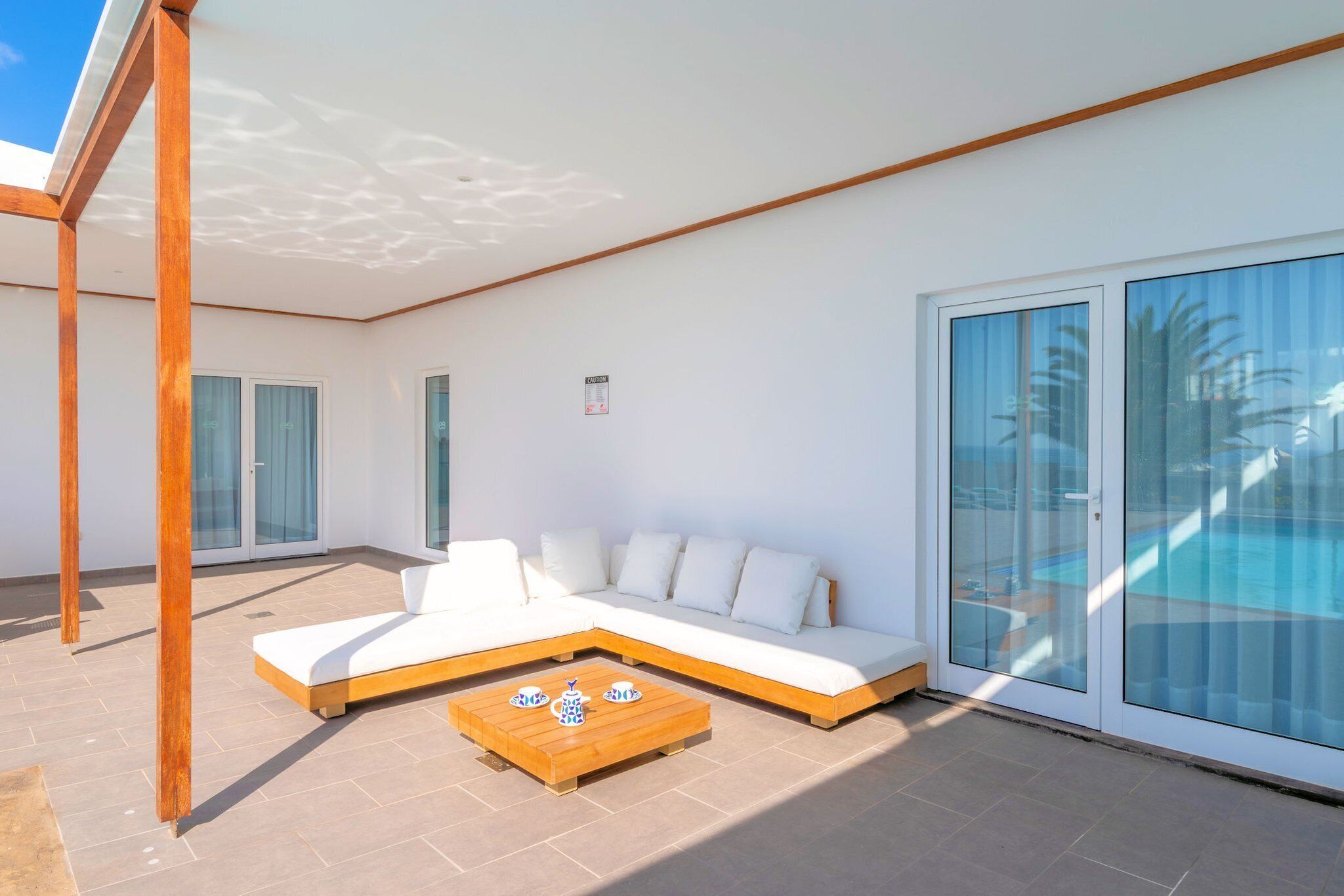 A modern outdoor living area with a sectional sofa, a wooden coffee table, and a glass door leading to a pool.