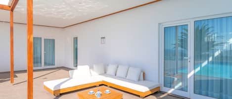 A modern outdoor living area with a sectional sofa, a wooden coffee table, and a glass door leading to a pool.