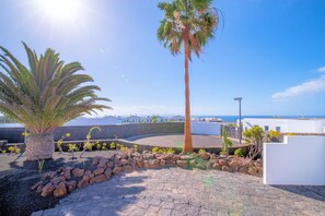 A coastal property with a palm tree, rocky garden, and a view of the sea and buildings in the distance. - Villa Maravento | Playa Blanca | Heated Pool | Family Luxury Retreat (Playa Blanca)