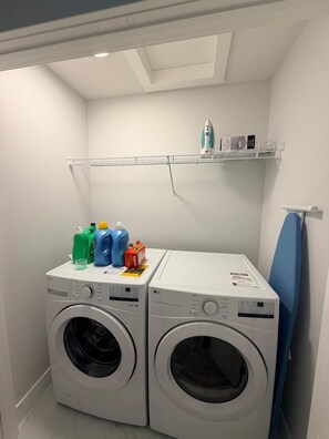 A laundry room with two front-loading washing machines, a drying rack, and a shelf with laundry supplies. - Modern Comfort Meets Cozy Charm | New 3BR Home in Chestermere, Alberta (Chestermere)