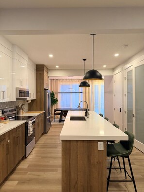 A modern kitchen with wooden cabinets, stainless steel appliances, a central island, and a pendant light. - Modern Comfort Meets Cozy Charm | New 3BR Home in Chestermere, Alberta (Chestermere)