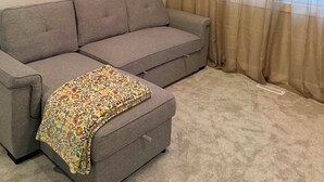 Ein Wohnzimmer mit einem grauen Sofa, einer gemusterten Decke und transparenten Vorhängen.