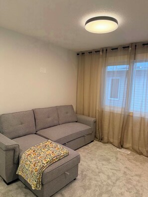 Ein Wohnzimmer mit einem grauen Sofa, einer gemusterten Decke und transparenten Vorhängen.