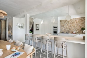 A modern kitchen with a stone backsplash, bar stools, and a dining area with a wooden table. - Villa Azurea | Almancil Algarve | 4BR Retreat | Private Pool & Terrace (Almancil)