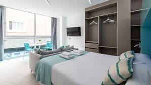 Un dormitorio moderno con una cama, un área de descanso con una mesa y sillas, un televisor montado en la pared y un ventanal con cortinas.