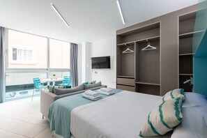 A modern bedroom with a bed, a seating area with a table and chairs, a TV mounted on the wall, and a large window with curtains. - Studio in Las Palmas Near Playa de Las Canteras (Las Palmas de Gran Canaria)